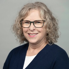 Professional headshot of a smiling individual wearing glasses and a navy blue cardigan.