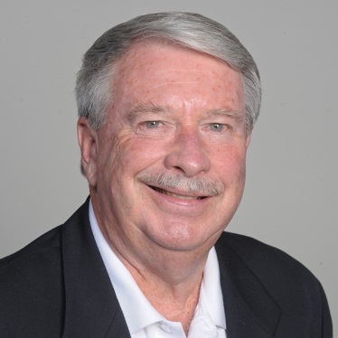 A person with short gray hair and a gray mustache, wearing a white collared shirt and dark blazer, smiling warmly against a neutral gray background.