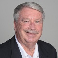A person with short gray hair and a gray mustache, wearing a white collared shirt and dark blazer, smiling warmly against a neutral gray background.