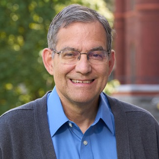 Portrait of a person smiling, wearing glasses and a blue shirt, with a blurred background of greenery and a building.