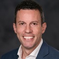 Portrait of a smiling person in a blue suit against a dark gray background.