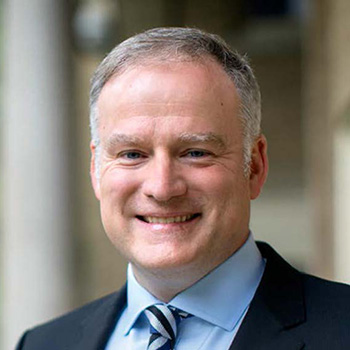 Portrait of a smiling individual in a business suit, standing outdoors with a building in the background.