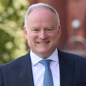 Portrait of a smiling individual in a business suit, standing outdoors with trees and a brick building in the background.