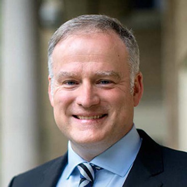 Portrait of a smiling individual in a business suit, standing outdoors with a building in the background.
