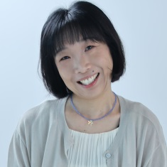 A person with short black hair and bangs, smiling, wearing a light gray cardigan over a cream top and a thin beaded necklace with a small star pendant, against a plain light blue background.