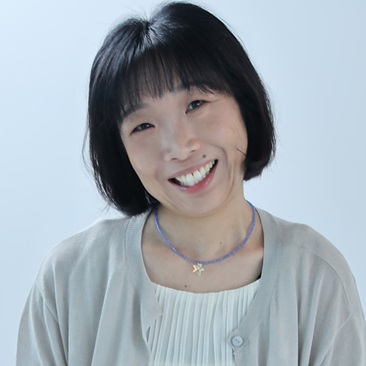 A person with short black hair and bangs, smiling, wearing a light gray cardigan over a cream top and a thin beaded necklace with a small star pendant, against a plain light blue background.