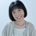 A person with short black hair and bangs, smiling, wearing a light gray cardigan over a cream top and a thin beaded necklace with a small star pendant, against a plain light blue background.