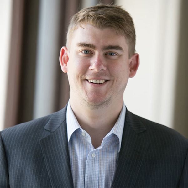 A person wearing a gray pinstripe blazer and a light blue checkered dress shirt, set against a softly blurred indoor background.