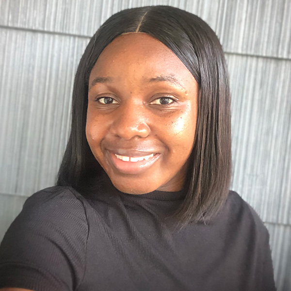 Close-up portrait of an individual smiling at the camera, wearing a black top, set against a textured gray background.
