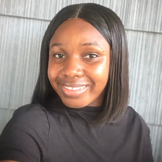 Close-up portrait of an individual smiling at the camera, wearing a black top, set against a textured gray background.