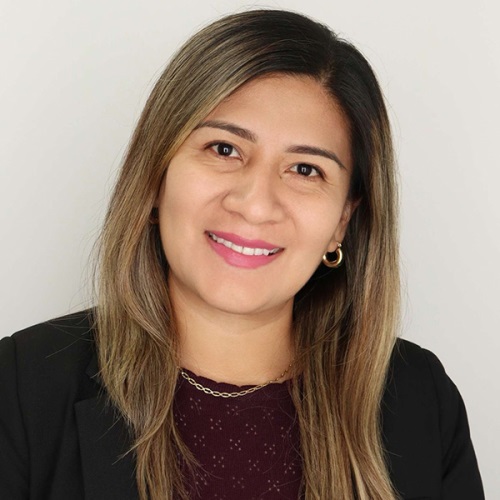 Professional headshot of a smiling individual with long hair, wearing a dark blouse and a gold necklace.