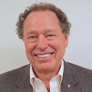 A person with curly gray hair smiles broadly, wearing a gray blazer and open-collar white shirt against a light gray background.