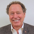 A person with curly gray hair smiles broadly, wearing a gray blazer and open-collar white shirt against a light gray background.