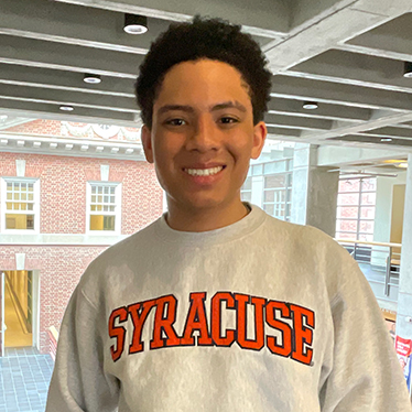 A person  smiles at the camera wearing a gray Syracuse University sweatshirt. A brick building is visible through large windows in the background.