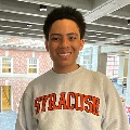 A person  smiles at the camera wearing a gray Syracuse University sweatshirt. A brick building is visible through large windows in the background.