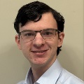 A person with dark hair and glasses weaing a collared shirt.