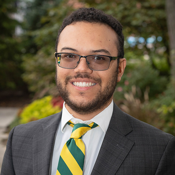 A person wearing eyeglasses and a suit with a green and yellow striped tie is smiling outdoors with greenery in the background.