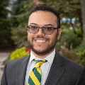A person wearing eyeglasses and a suit with a green and yellow striped tie is smiling outdoors with greenery in the background.
