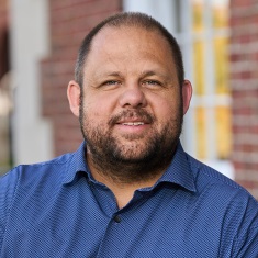 Portrait of a smiling person in a blue shirt, standing in front of a blurred brick building background.