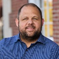 Portrait of a smiling person in a blue shirt, standing in front of a blurred brick building background.