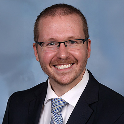 A person wearing glasses, a dark suit and a white shirt and striped tie.