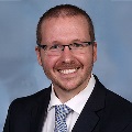 A person wearing glasses, a dark suit and a white shirt and striped tie.