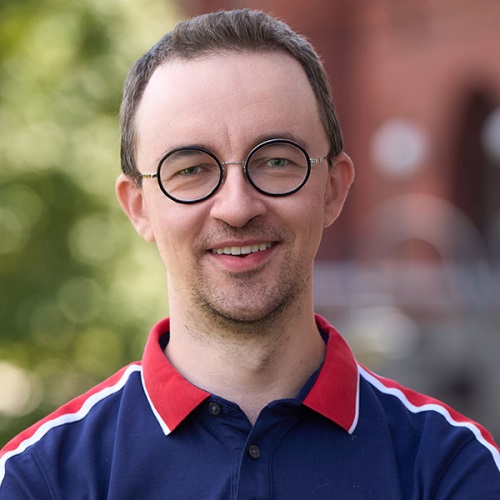 Portrait of an individual wearing round glasses and a polo shirt, smiling, with a blurred background of red brick buildings.
