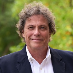 Headshot of a smiling person with curly hair, wearing a dark blazer over a light-colored shirt, set against a blurred natural background.