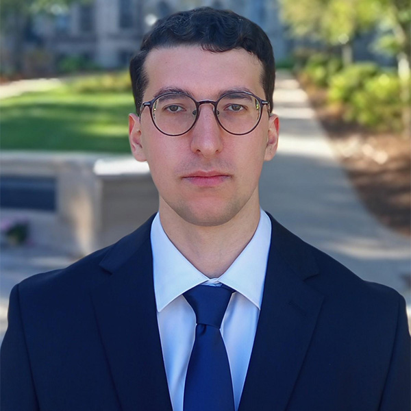 A person in a navy suit and tie, wearing glasses, standing outdoors on a campus path.