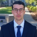 A person in a navy suit and tie, wearing glasses, standing outdoors on a campus path.