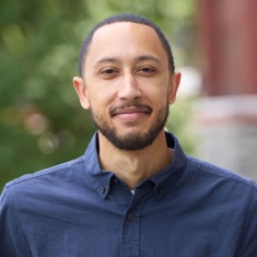 Portrait of an individual in a blue shirt, smiling and standing outdoors.