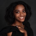 Portrait of a smiling person with curly hair wearing a black top against a dark background.