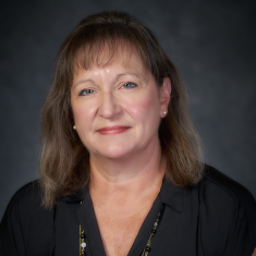 Professional headshot of an individual with shoulder-length brown hair, wearing a black shirt and a necklace, against a grey background.