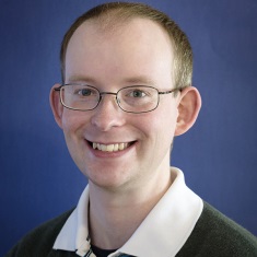 miling, wearing wire-rimmed glasses and a dark polo shirt, against a blue background.