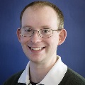 miling, wearing wire-rimmed glasses and a dark polo shirt, against a blue background.
