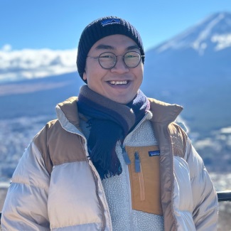 Photo of a smiling person wearing glasses, a scarf and a winter coat with snowy mountains in the background.