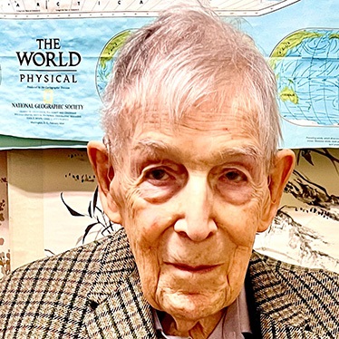 An elderly person posing in front of a National Geographic map titled 'The World Physical.'