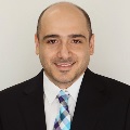 A person smiling in dark suit with blue and purple plaid tie.