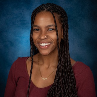 Portrait of a smiling person with braided hair, wearing a maroon top and a necklace against a blue background.