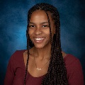 Portrait of a smiling person with braided hair, wearing a maroon top and a necklace against a blue background.