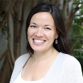 A person smiling in light blazer, feather earrings, trees in background.