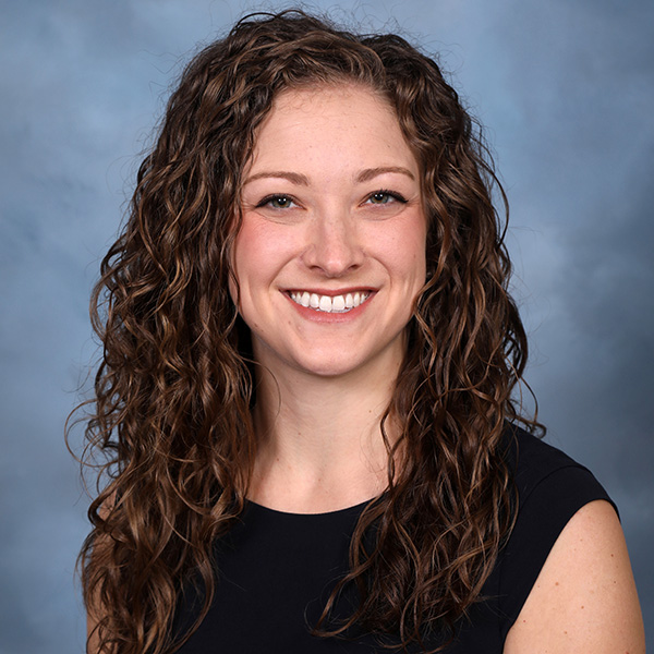 A person smiling with long curly brown hair and wearing a dark sleeveless top, set against a soft blue-gray background