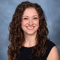 A person smiling with long curly brown hair and wearing a dark sleeveless top, set against a soft blue-gray background