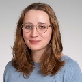 Portrait of a person with shoulder-length hair wearing round glasses and a blue shirt, against a light gray background.