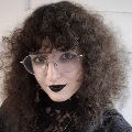 Portrait of a person with dark curly hair and glasses wearing a black top.
