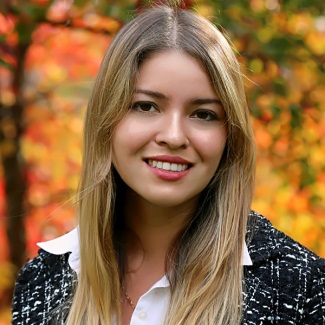 A person with long blonde hair, smiling and wearing a black and white top with blurry fall trees in the background.
