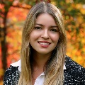 A person with long blonde hair, smiling and wearing a black and white top with blurry fall trees in the background.