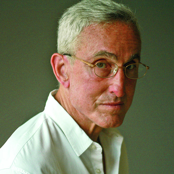A person with short gray hair and gold-framed round glasses looks toward the camera against a neutral gray background. They are wearing a white collared shirt.