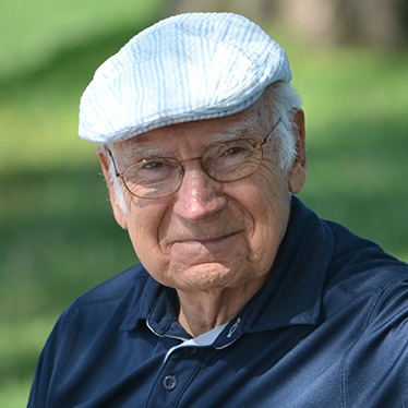 Close-up portrait of an elderly person wearing a striped cap and a blue polo shirt, smiling gently in a park setting.