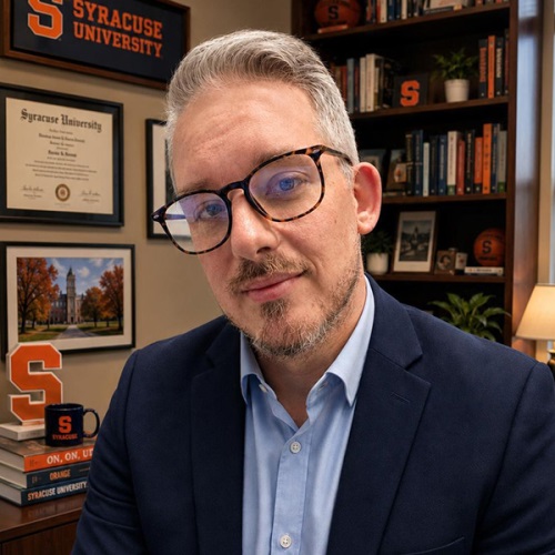 A person with short gray hair and a beard, wearing tortoiseshell glasses and a navy blazer over a light blue dress shirt. They are seated in an office decorated with Syracuse University memorabilia